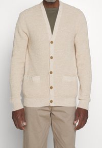 Knit cardigan in light beige with a V-neck, five buttons, two front pockets, and ribbed cuffs. Worn over a dark green shirt.