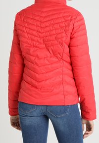 Rød dunjakke med chevron-quiltet mønster, høj krave og elastiske manchetter. Bæres med blå denimjeans, der fremhæver et tætsiddende design.