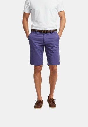 Uomo che indossa pantaloncini viola fino al ginocchio, t-shirt bianca a maniche corte, cintura marrone intrecciata e scarpe marroni con lacci, in piedi con le mani nelle tasche.