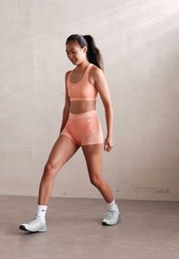 Lyse ferskenfarget sports-bh og matchende shorts, Nike-logo på begge, kombinert med lyseblå treningssko og hvite sokker. Glatt tekstur.