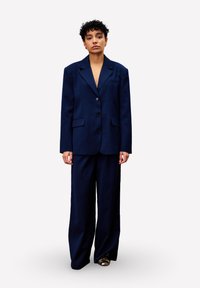 Persoon met kort krullend haar, gekleed in een losse donkerblauwe pak en metallic schoenen, rechtop staand tegen een effen achtergrond.