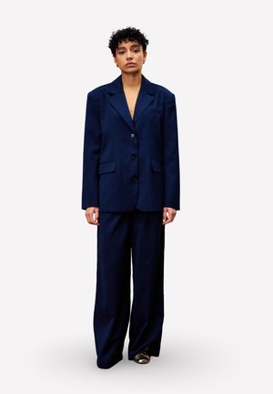 Persoon met kort krullend haar, gekleed in een losse donkerblauwe pak en metallic schoenen, rechtop staand tegen een effen achtergrond.