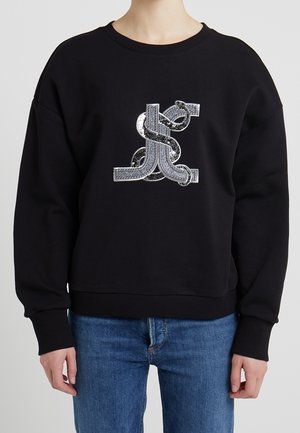 Sweatshirt noir avec un design graphique en sequins argentés texturés. Coupe décontractée avec des poignets et un ourlet côtelés. Porté avec un jean bleu.