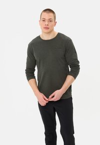 Camicetta a maniche lunghe di colore verde oliva con scollo rotondo, tessuto morbido e dettagli del logo discreti sul petto. Abbinata a pantaloni neri.