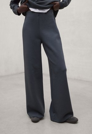 Persona con pantalones anchos de pierna oscuros y zapatos a juego, levantando un jersey gris para revelar una camisa blanca debajo.