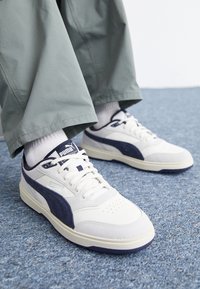 Sneakers bianchi con accenti in camoscio blu navy, tomaia in pelle texturizzata, perforazioni sulla punta, lacci bianchi e suola in gomma resistente. Design casual e a profilo basso.
