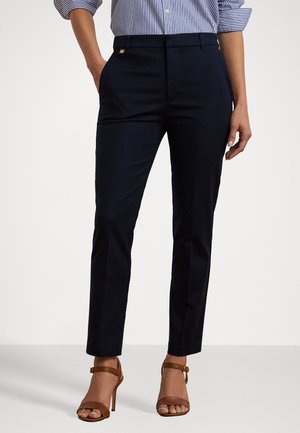 Femme portant un pantalon noir slim et des sandales à talons hauts marron, associée à une chemise rayée bleue et blanche avec les manches retroussées.