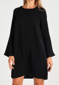Svart långärmad klänning med rund halsringning och utställda ärmslut. Tillverkad av slät tyg, med en A-linjeform och knä-till ovan längd.