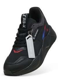 Fekete sportcipő hálós és bőr anyagokkal, piros és kék részletekkel, vastag talppal, hangsúlyos logóval és ezüst címkével.