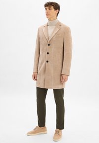Beige Cordjacke mit Reverskragen und schwarzen Knöpfen, kombiniert mit olivgrünen Hosen und beigen Schuhen. Glatter Stoff und strukturierte Passform.