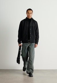 Casaco preto de botões sobre um gola alta preto, combinado com calças de ganga verde largas e sapatos pretos, segurando uma mala preta de ombro.