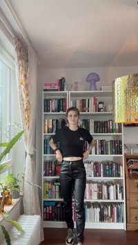 T-shirt noir court, pantalon en cuir noir taille haute et baskets multicolores. Salle lumineuse avec des étagères à livres et des plantes décoratives.