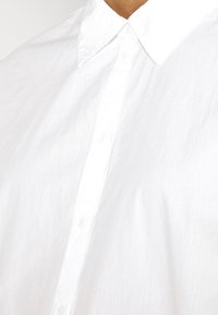 Bílá košile s knoflíky vyrobená z lehké tkaniny, s klasickým límcem a průhlednými plastovými knoflíky podél předního zapínání.