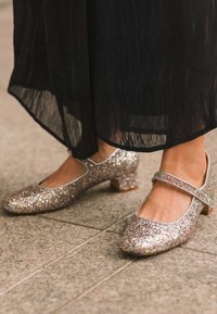 Zapatos Mary Jane plateados brillantes con tacón bajo, punta redondeada y detalles metálicos, combinados con una prenda negra con plisados, sobre una superficie de piedra.