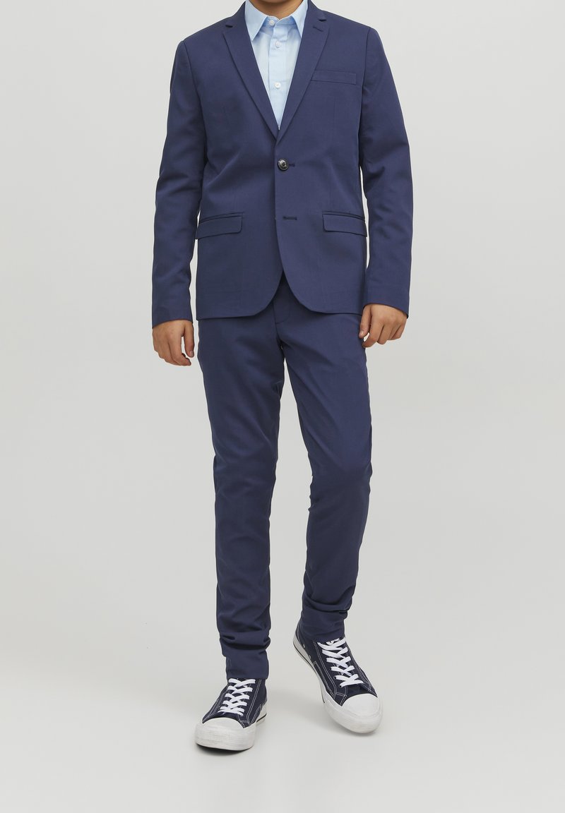 Enfant portant un costume bleu marine avec une chemise bleu clair et des baskets montantes bleu marine, debout devant un fond clair uni.
