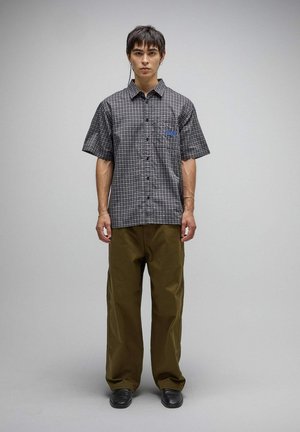 Giovane uomo in piedi dritto con camicia a maniche corte a quadri bianco e nero, pantaloni larghi verde oliva e scarpe nere su sfondo grigio uniforme.