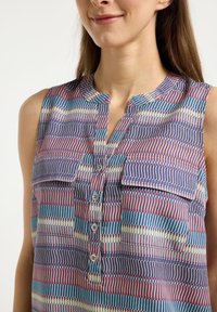 Camicetta senza maniche con bottoni, caratterizzata da un motivo geometrico multicolore in blu navy, rosa e teal, con due taschini sul petto e scollatura rotonda.