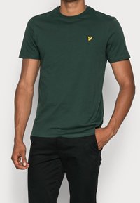 Grünes Baumwoll-T-Shirt mit klassischem Rundhalsausschnitt und kurzen Ärmeln. Mit einem kleinen gelben gestickten Logo auf der linken Brust.