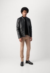 Geacă din piele neagră cu guler înalt, purtată cu o cămașă neagră, pantaloni de cânepă bej și cizme negre până la gleznă, stând pe un fundal neutru.