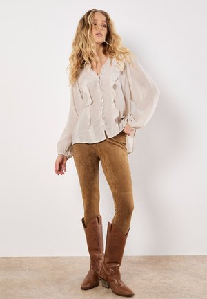 Blonde vrouw in een lichtbeige rimpelblouse met knopen, bruine suède aangesloten broek en lange bruine leren laarzen, poseert binnen.