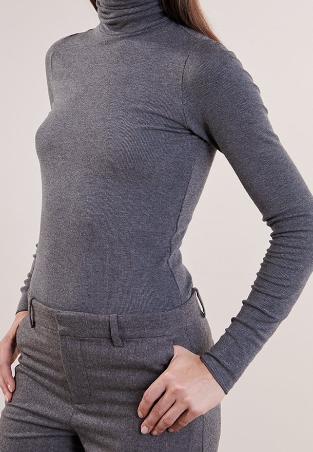 Femme portant un pull gris à col roulé ajusté et un pantalon gris assorti, avec une main posée sur la hanche.