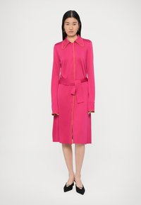 Różowa sukienka zapinana na guziki z długimi rękawami, z paskiem w talii, żółte wykończenie wzdłuż kołnierzyka i listwy guzika, o długości do kolan z rozcięciem po boku. Czarne buty.