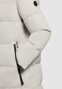 Giacca puffer grigia chiara con tasca con zip, texture morbida e logo nero rotondo sulla manica. Cucitura dettagliata visibile.
