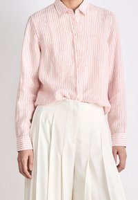 Jasnoróżowa, paskowana koszula z guzikami, wykonana z miękkiego materiału, z klasycznym kołnierzykiem, długimi rękawami i białymi guzikami. Noszona z plisowaną białą spódnicą.