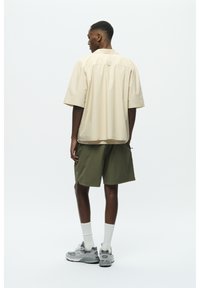 Beige Kurzarmhemd, lockere Passform, kombiniert mit olivgrünen Shorts. Das Model trägt graue Turnschuhe und weiße Socken. Einfaches Design, keine Muster.
