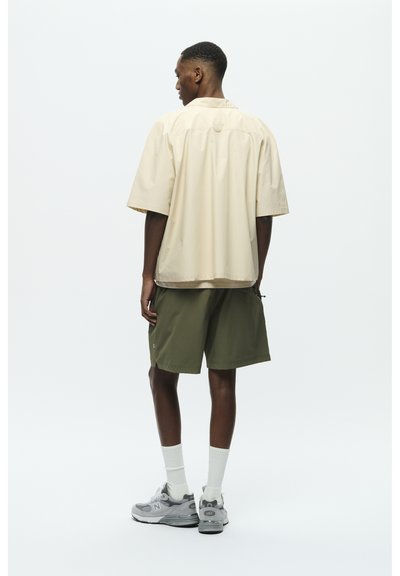 Chemise beige à manches courtes, coupe décontractée, portée avec un short vert olive. Le modèle porte des baskets grises et des chaussettes blanches. Design simple, sans motifs.