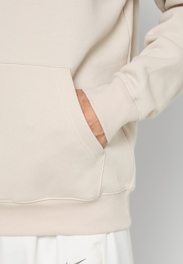 Person trägt einen beigen Hoodie mit einer Hand teilweise in der vorderen Tasche, kombiniert mit weißen Nike-Shorts, die ein graues Swoosh-Logo zeigen.