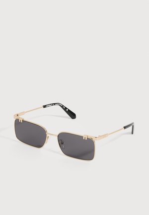 Lunettes de soleil rectangulaires avec verres noirs, monture fine en métal doré et embouts de branches noirs, inclinées sur un fond gris clair.