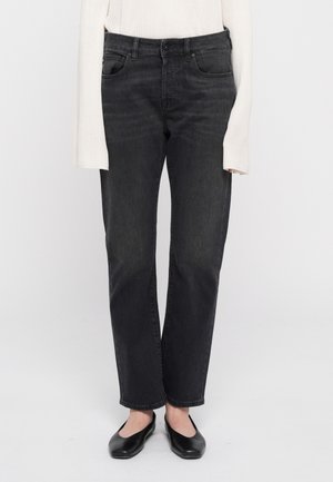 Personne portant un jean noir droit, un pull blanc à manches longues côtelé et des chaussures plates noires, debout devant un fond blanc.