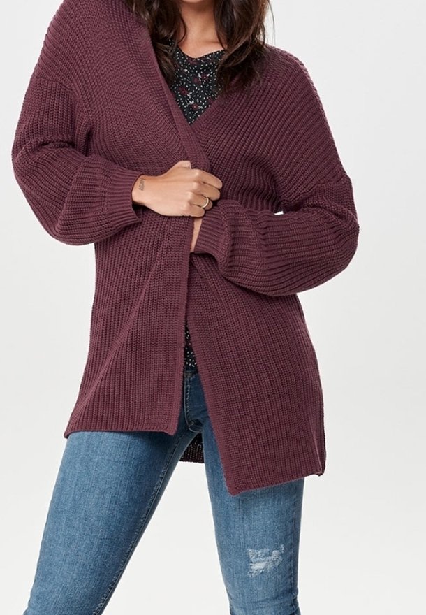 Gebreide cardigan in bordeauxrood, met een open voorkant en ribbelstructuur, gecombineerd met een zwart bloemenbloesje en blauwe versleten jeans.