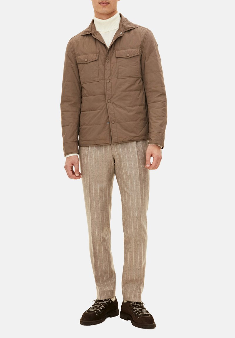 Giacca trapuntata marrone con due tasche frontali, abbinata a pantaloni beige a righe e scarpe marrone scuro. Modello che indossa un dolcevita bianco.