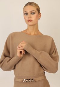 Beige gerippter Pullover mit weiten Ärmeln und rundem Halsausschnitt, mit einem Gürtel, der eine metallische Kettenakzent um die Taille hat.