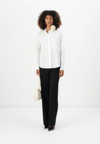 Camicia bianca con bottone e tasca sul petto, abbinata a pantaloni neri su misura. Accessoriata con una borsa color crema e dettagli floreali.