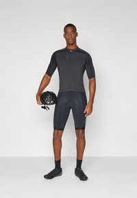 Maglietta da ciclismo nera a maniche corte con zip frontale, abbinata a pantaloni corti neri aderenti. Il ciclista tiene in mano un casco nero.