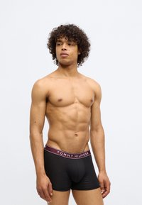 Shirtless man with curly hair wearing black Tommy Hilfiger boxer briefs, standing against a plain light background, looking left.