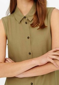 Camicia senza maniche verde oliva con bottoni indossata da una persona con le braccia incrociate e capelli castani lunghi fino alle spalle, mostrata dalla zona del collo fino alla vita.