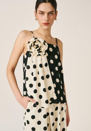 Mujer con blusa sin mangas de lunares blancos y negros con una gran flor de tela y pantalones a juego con lunares, de pie con una mano en el bolsillo.