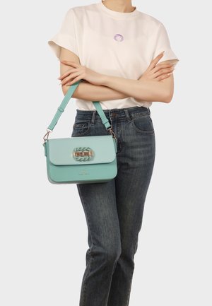 Donna che indossa una maglietta bianca e jeans scuri, che tiene con le braccia incrociate una borsa a tracolla azzurro chiaro con fibbia dorata e cinturino turchese.