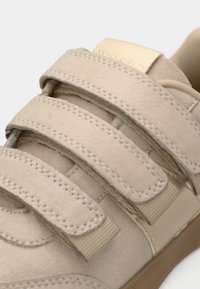 Baskets en daim beige avec trois bandes Velcro, des détails cousus, un embout texturé et un logo doré sur la languette.