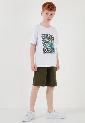 Red-haired boy wearing white T-shirt with colorful Tyrannosaurus Rex graphic, olive green shorts, and white sneakers, standing on white background.
