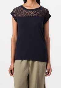 Marineblå T-shirt med et transparent mønster i spidslace i den øverste del, rund halsudskæring og korte optrillede ærmer; parret med lyse strukturerede vidde bukser.