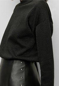 Maglione nero a maglia con collo alto e spalle cadenti, abbinato a una gonna in pelle nera con borchie a forma di piramide argentate lungo la cucitura.
