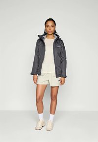 Grå vattentät jacka med huva, fram zip, och logotyp patch, kombinerad med en beige topp och shorts, samt ljusa sneakers.