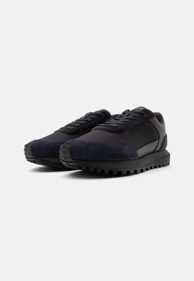 Paire de baskets basses noires avec semelles texturées, design à lacets et détails en daim sur les orteils et les talons sur fond blanc uni.