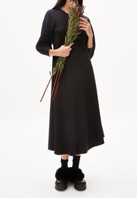 Zwarte jurk tot de knieën met lange mouwen en een ronde hals. Model houdt groen plantenmateriaal vast en draagt zwarte fuzzy schoenen met donkere sokken.