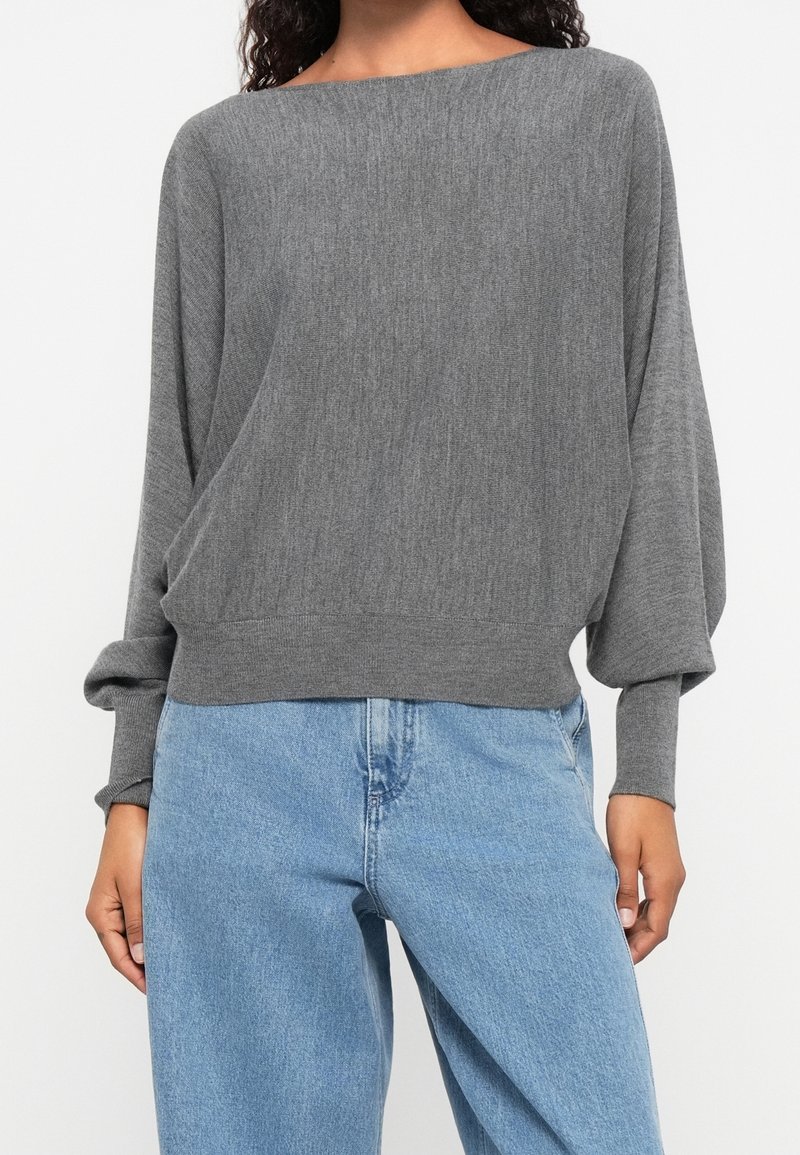 Personne portant un pull gris ample à manches longues avec des manches bouffantes et un jean bleu clair taille haute, debout.